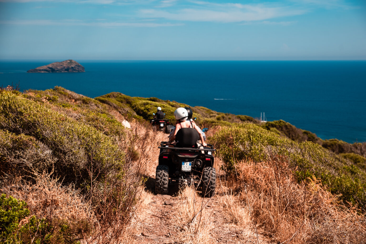 Quad in fila con pilota e passeggero col casco su uno sterrato fra la macchia, con il mare del sud-ovest della Sardegna